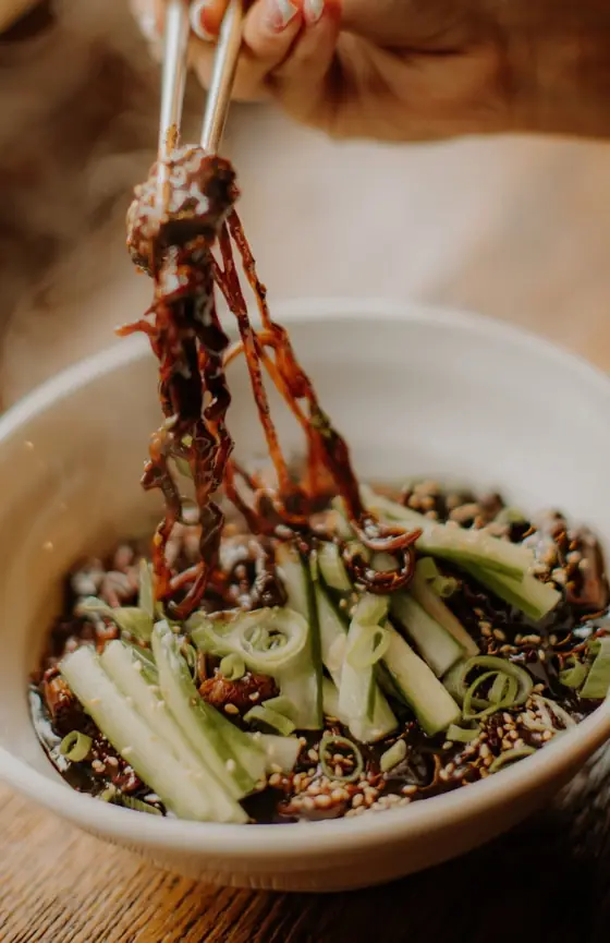 Makaron jajangmyeon w ciemnym sosie podnoszony pałeczkami nad miską z ogórkiem